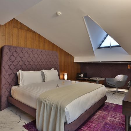 Dosso Dossi Hotels & Spa Downtown Istambul Exterior foto The photo shows a stylish hotel room with a modern design. It features a large bed with a tufted purple headboard, complemented by white bedding and a light gray throw. The walls are made of wooden panels, adding warmth to the space. To the side, the