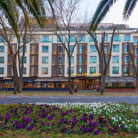 Dosso Dossi Hotels & Spa Downtown Istambul Exterior foto The photo shows a building with a modern design, featuring a facade with large windows and a mix of materials. In the foreground, there are colorful flower beds with purple and white flowers. The scene is framed by tall trees, and the overall setting
