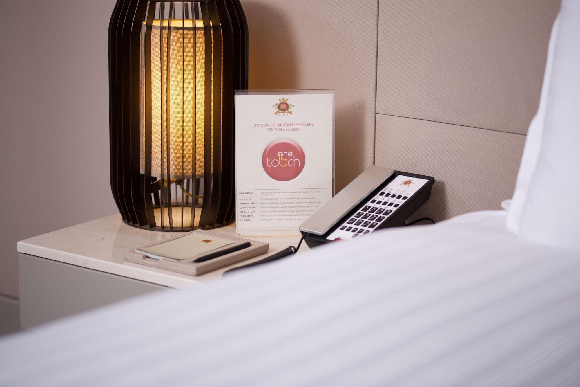 Dosso Dossi Hotels & Spa Downtown Istambul Exterior foto The photo shows a bedside setup in a room. There is a stylish lamp with a warm light on a table, alongside a menu or information card that features a logo or brand name. A remote control is also placed on the table, suggesting it might be used for va
