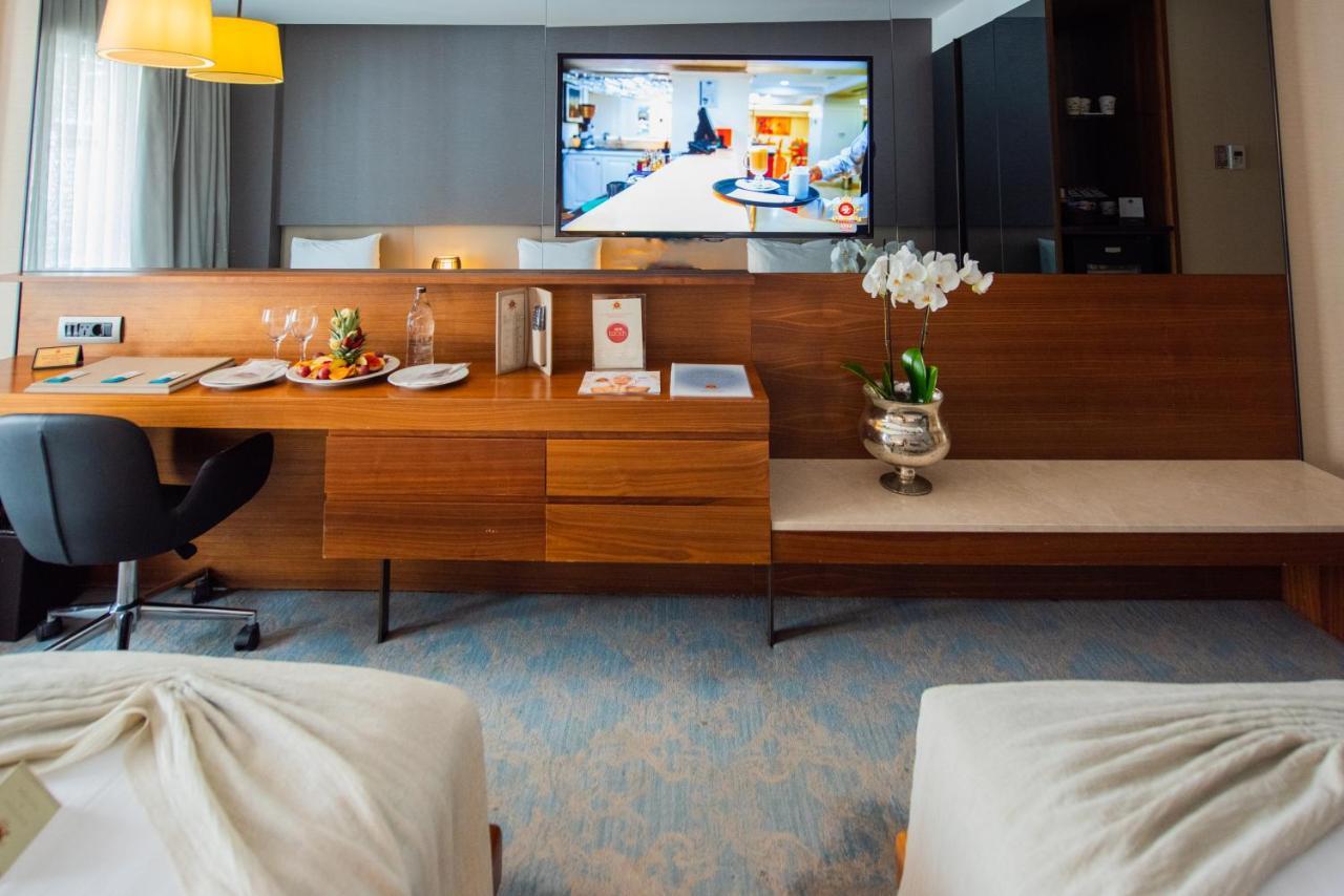 Dosso Dossi Hotels & Spa Downtown Istambul Exterior foto The photo shows a modern hotel room featuring two beds with light-colored bedding. In the background, there is a flat-screen television mounted on the wall displaying what appears to be a hotel service or entertainment program. A wooden console table