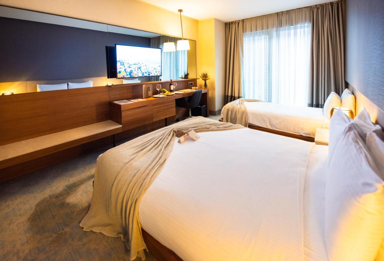 Dosso Dossi Hotels & Spa Downtown Istambul Exterior foto The photo shows a modern hotel room featuring two beds with white linens and a decorative throw blanket. There's a wooden desk and a chair alongside a large flat-screen TV mounted on the wall. The room has large windows with curtains, allowing natura