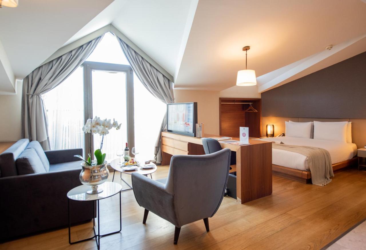 Dosso Dossi Hotels & Spa Downtown Istambul Exterior foto The photo shows a stylish hotel room with a modern design. It features a comfortable bed in one area, complemented by a cozy seating area with a sofa and a chair. There is a small round table with a vase of flowers and some snacks. A flat-screen TV i