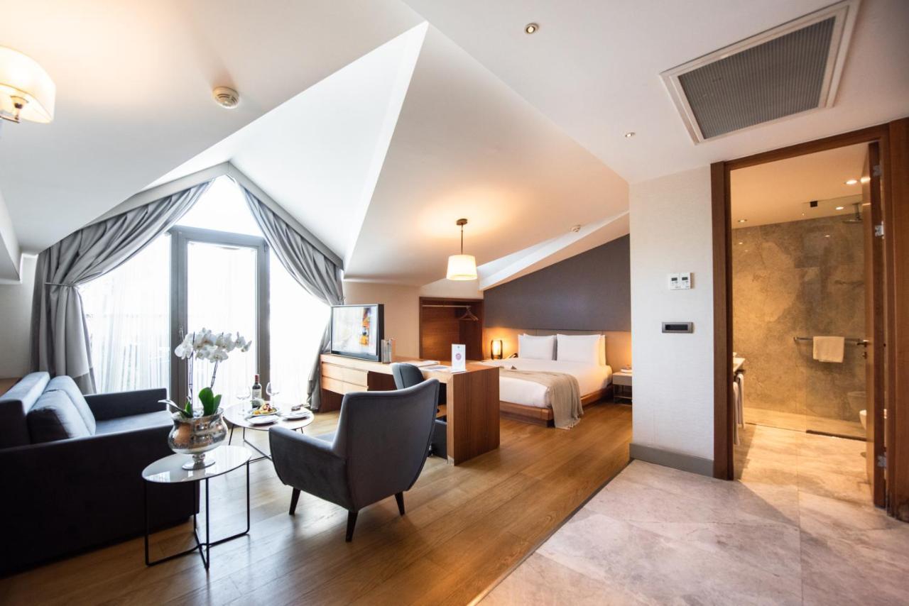 Dosso Dossi Hotels & Spa Downtown Istambul Exterior foto The photo shows a stylish hotel room with a modern design. It features slanted ceilings and large windows that allow natural light to flood the space. The room includes a comfortable seating area with a sofa and a small table, as well as a well-made 