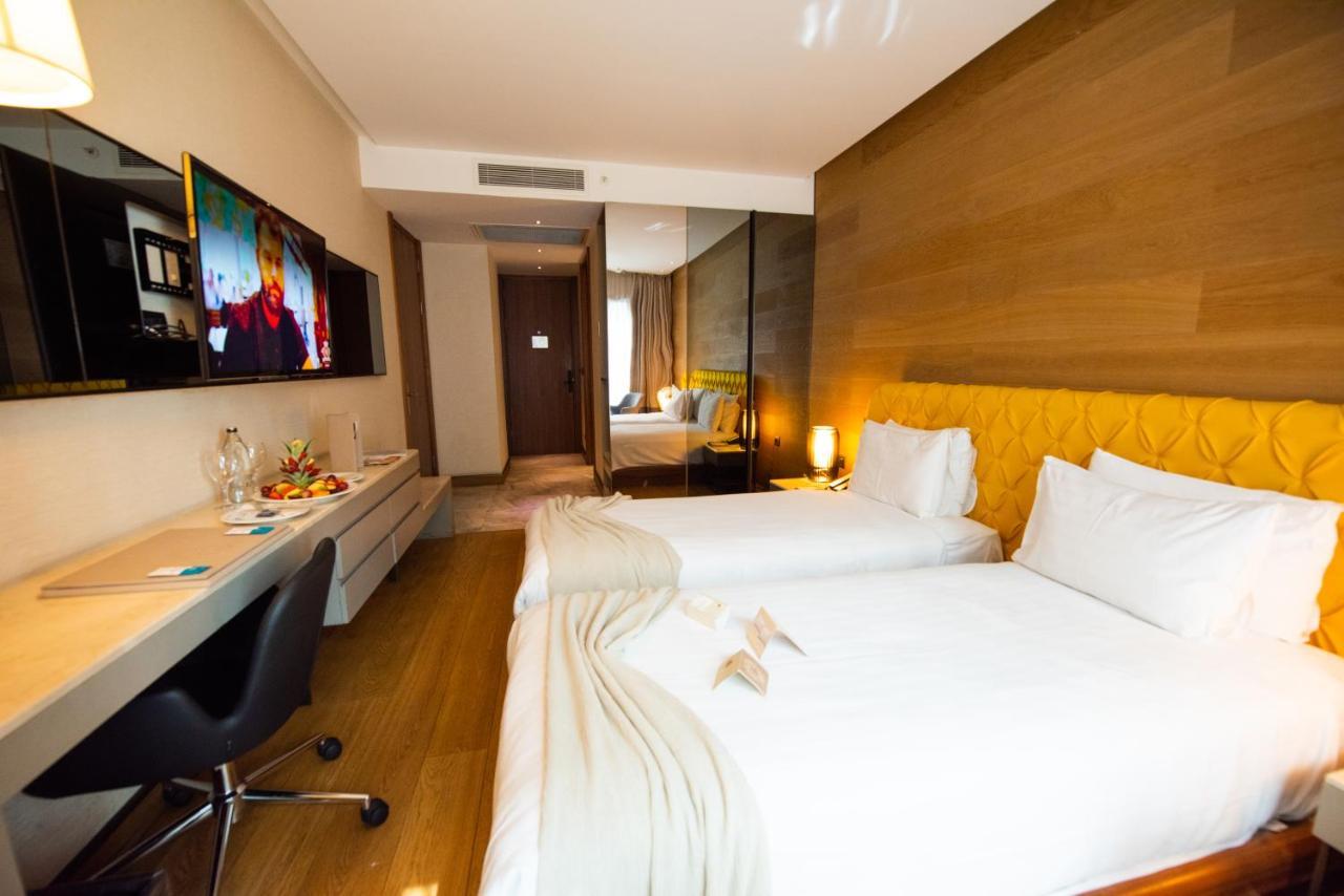 Dosso Dossi Hotels & Spa Downtown Istambul Exterior foto The photo shows a modern hotel room featuring two single beds with white linens. There is a stylish yellow accent wall behind the beds. A flat-screen television is mounted on the wall opposite the beds, and a sleek desk and chair are positioned next 