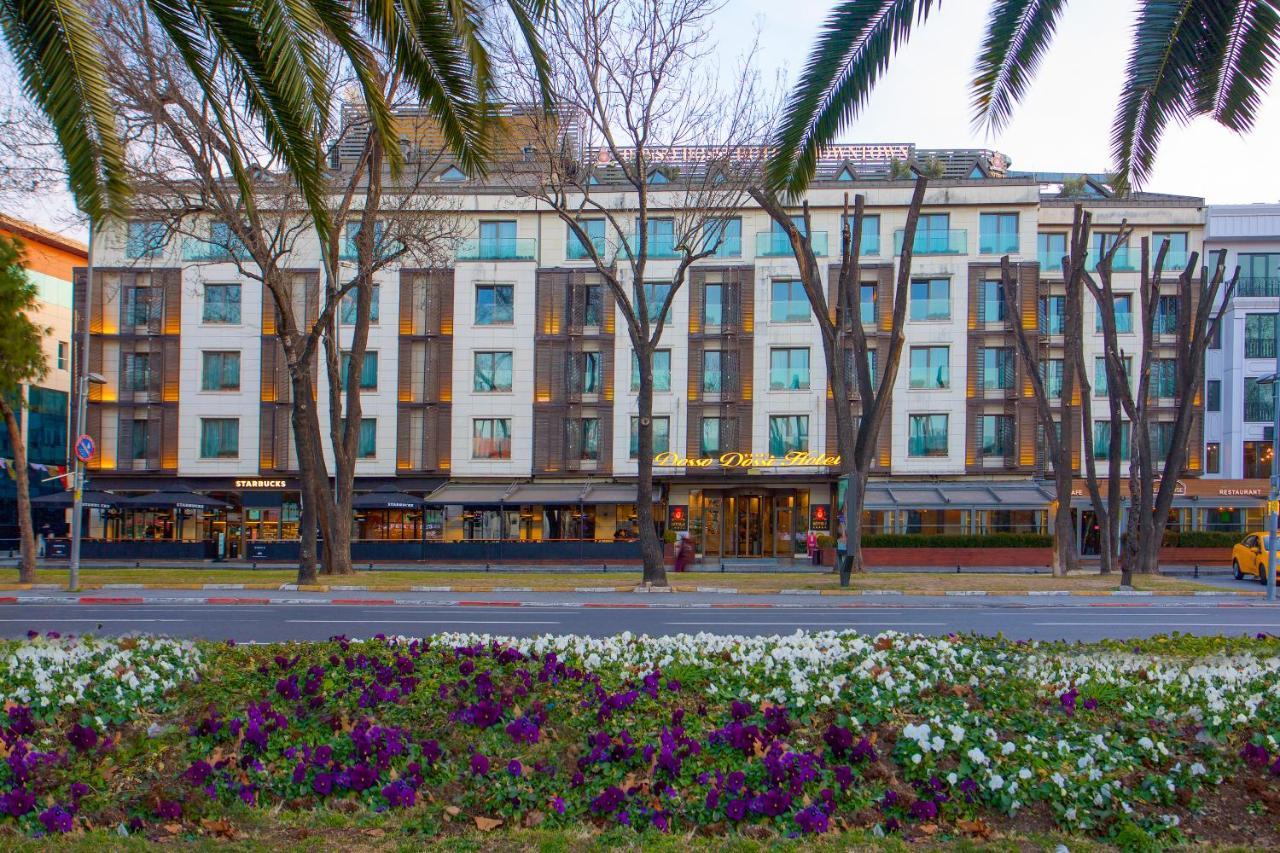 Dosso Dossi Hotels & Spa Downtown Istambul Exterior foto The photo shows a building with a modern design, featuring a facade with large windows and a mix of materials. In the foreground, there are colorful flower beds with purple and white flowers. The scene is framed by tall trees, and the overall setting