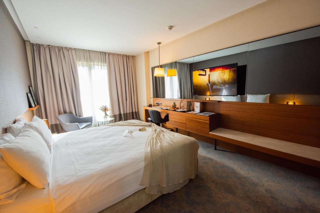Dosso Dossi Hotels & Spa Downtown Istambul Exterior foto The photo shows a modern hotel room featuring a large bed with white linens, set against a soft-toned wall. There is a stylish gray armchair in one corner and a sleek wooden desk with a chair. The room is well-lit, with large windows covered by light