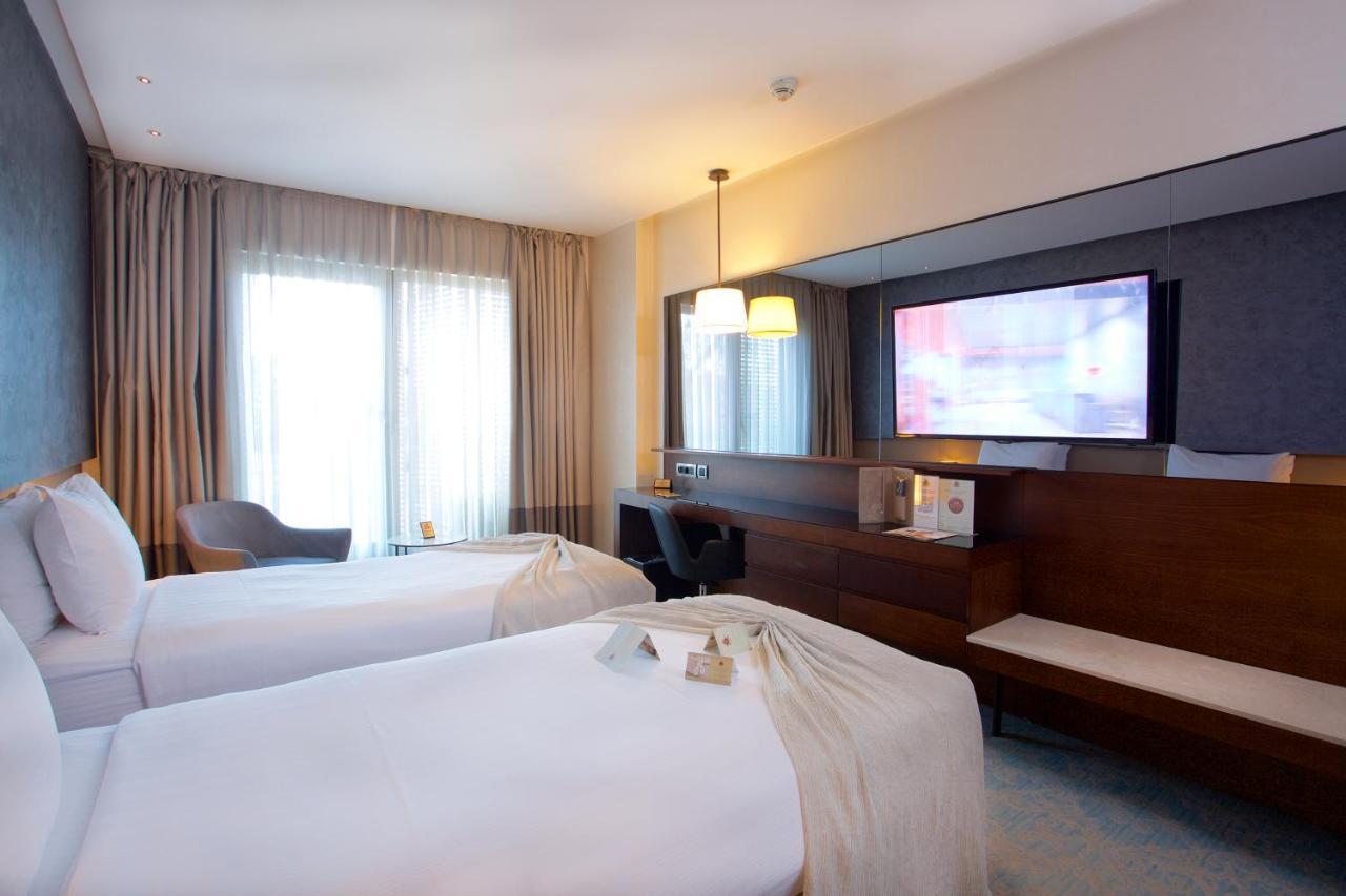 Dosso Dossi Hotels & Spa Downtown Istambul Exterior foto The photo shows a hotel room featuring two twin beds with white linens. There is a large window with curtains that allows natural light to enter the room. A wooden cabinet is present, along with a flat-screen television mounted on the wall. Additiona