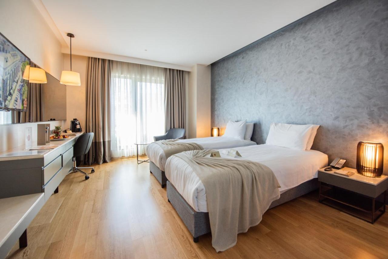 Dosso Dossi Hotels & Spa Downtown Istambul Exterior foto The photo shows a modern hotel room featuring two single beds with white linens. The room has a muted color palette, including a gray wall and light wooden flooring. Large windows allow natural light to flood the space, and there are sheer curtains f