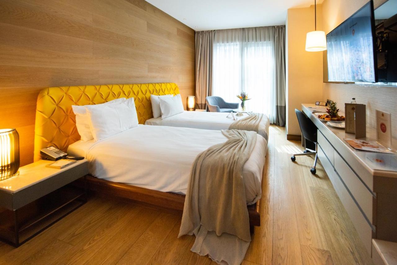 Dosso Dossi Hotels & Spa Downtown Istambul Exterior foto The photo shows a stylish hotel room featuring two beds with white linens and a unique yellow headboard. The room has wooden walls and a polished wooden floor. There are large windows with sheer curtains allowing natural light to flood in. A small se