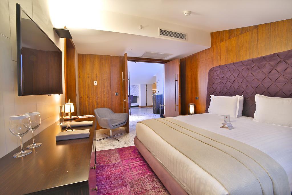 Dosso Dossi Hotels & Spa Downtown Istambul Exterior foto The photo depicts a modern hotel room. It features a large bed with white linens and a decorative headboard in a rich purple hue. The room has wooden paneling on the walls, adding warmth to the space. There's a flat-screen TV mounted on the wall oppo